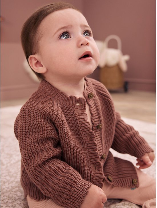 Baby met blauwe ogen in een bruin gebreid vest en lichtroze broek.