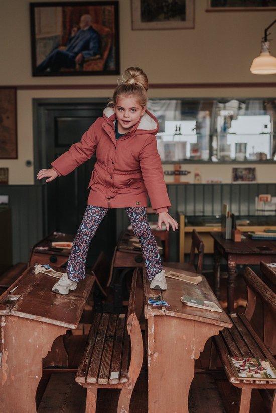 Een meisje met een roze winterjas en gebloemde flared broek staat op schoolbankjes.