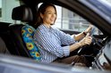 Vrouw in gestreepte blouse rijdt auto met blauw, bloemenkussen op stoel.