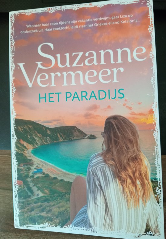Boek "Het Paradijs" van Suzanne Vermeer. Een vrouw kijkt uit over een Griekse baai met turquoise water en rotsachtige kust.