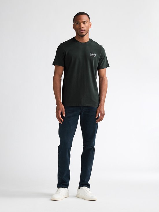 Man in donkergroen T-shirt met Petrol Industries logo op linkerborst. Draagt jeans en witte sneakers.