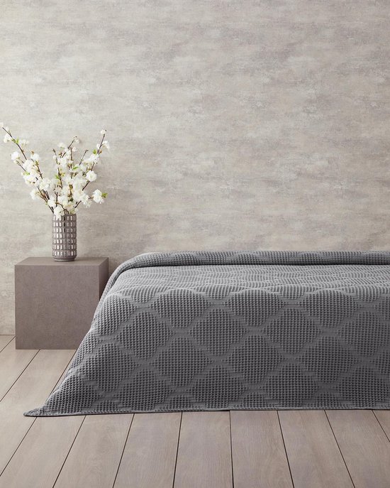 Grijze wafelsprei met cirkelmotief op een bed. Naast het bed staat een grijze bijzettafel met een vaas witte bloesems.