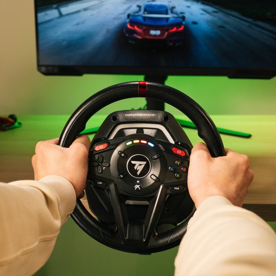 Handen van een persoon die een zwart racestuur vasthoudt met rode markering, voor een computerscherm.