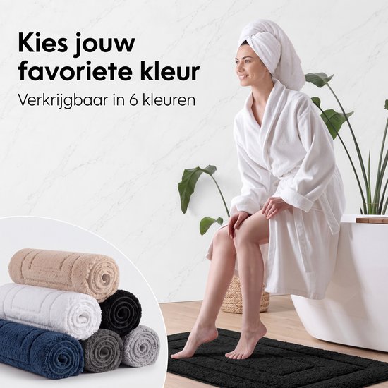 Vrouwen in badjas en met handdoek om haar hoofd, zittend op badrand. Duidelijke tekst 'Kies jouw favoriete kleur'. Gestapelde badmatten in verschillende kleuren: beige, wit, zwart, donkerblauw, grijs. Zwarte badmat op de vloer.