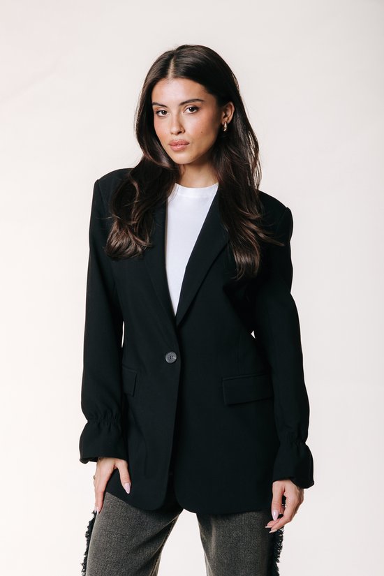 Model van voren gezien in een zwarte blazer, witte top en grijze broek.