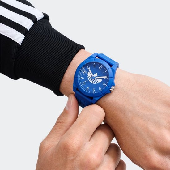 Close-up van een blauw Adidas horloge om een pols, gedragen met een zwart trainingsjack met witte strepen.