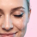 Vrouw met eyeliner en wimpers, met waterdruppels op haar gezicht. Waterbestendig product.