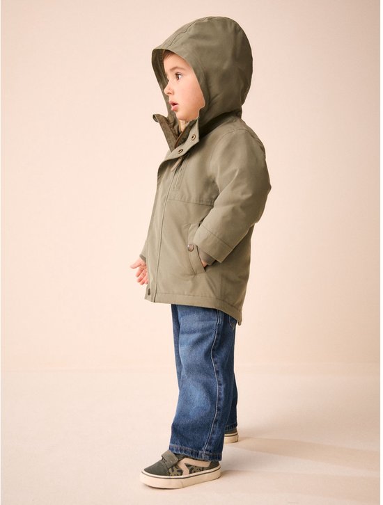 Zijaanzicht van een jongetje in een groene parka met capuchon, spijkerbroek en sneakers.