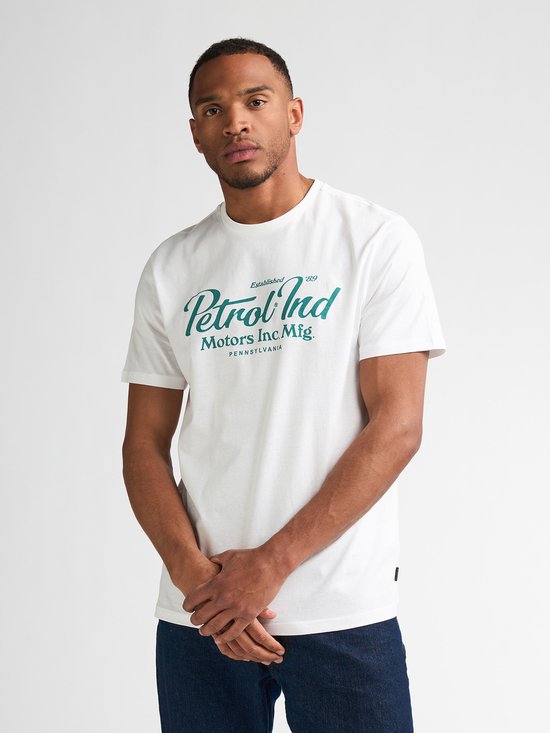 Man met donker haar, draagt wit T-shirt met groenblauwe print en spijkerbroek.