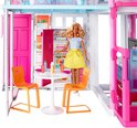 Barbie met gele rok en blauw-wit gestreept shirt in een keuken van een Barbiehuis, naast een tafel met twee oranje stoelen.