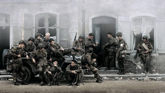 Groep militairen in uniformen en helmen, sommigen zittend op een jeep voor een gebouw.