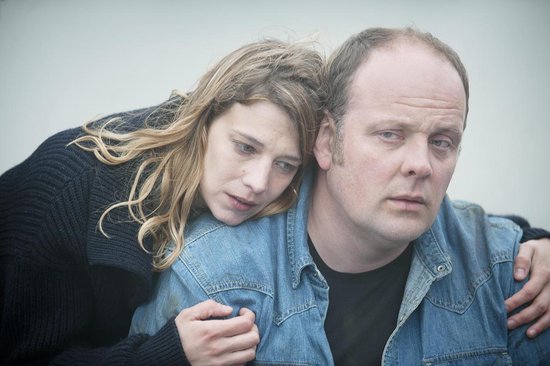 Een vrouw met blond haar leunt op de schouder van een man in een spijkerjack. Ze kijken allebei bezorgd weg.