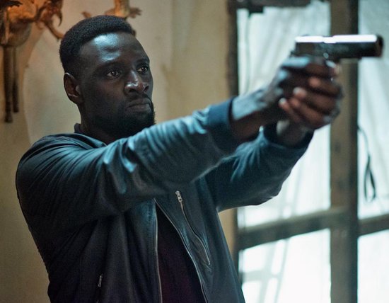 Omar Sy met donkere jas, gericht met een pistool, in een huiselijke omgeving.