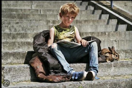 Jongen met lichtbruin haar zit op een trap met een deken en leest een boek, met een konijn naast hem.