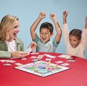 Een vrouw en twee kinderen spelen Monopoly Junior aan een rode tafel, met hun armen in de lucht.
