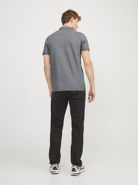 Man met kort bruin haar draagt een grijs gemêleerd poloshirt en zwarte jeans.