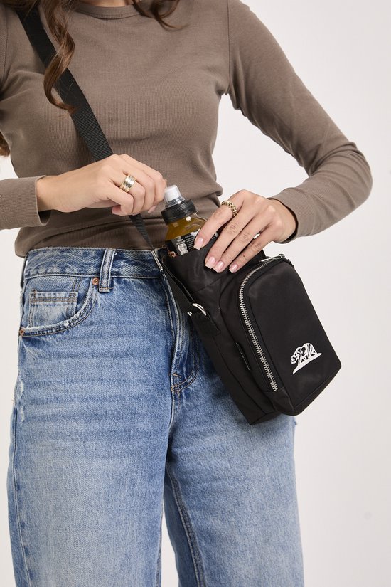 Vrouw in spijkerbroek en bruine longsleeve plaatst een flesje in een zwarte crossbody tas.