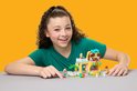 Une fille aux cheveux bouclés sourit et pointe du doigt une animalerie LEGO Friends. La boutique a une grosse tête de chien comme toit.
