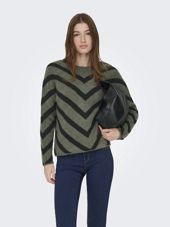 Model met olijfgroene trui met zwarte V-strepen, spijkerbroek en zwarte tas.