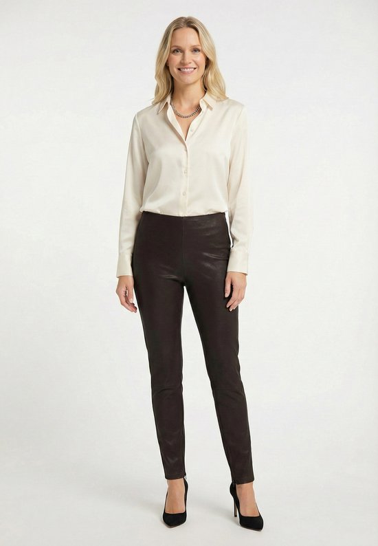 Vrouw met blond haar, voorkant, draagt een beige blouse en een donkerbruine leren broek.