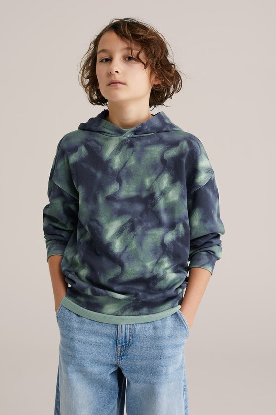 Jongen in een donkerblauwe en groene tie-dye hoodie en spijkerbroek.