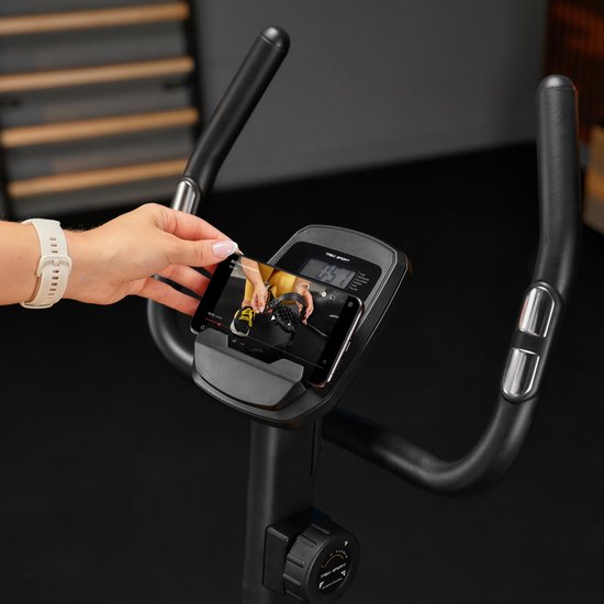 Close-up van een hand die een smartphone op het display van een hometrainer plaatst.