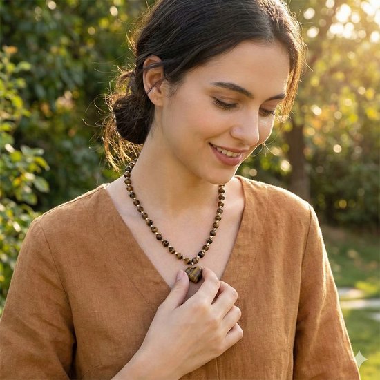 Model draagt een bruin-zwarte kralenketting met hartvormige tijgeroog hanger en gouden accenten, in de natuur.