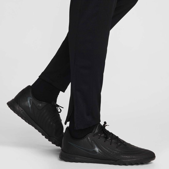 Detail van de onderkant van een zwarte Nike trainingsbroek met rits aan de zijkant en zwarte schoenen.