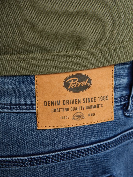 Close-up van het bruine leren 'Petrol' label op een blauwe denim short. Het label vermeldt 'DENIM DRIVEN SINCE 1989 CRAFTING QUALITY GARMENTS'.