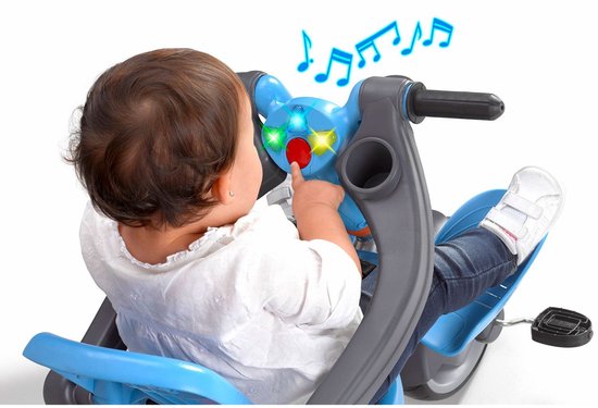 Baby zit op een blauwe driewieler met oranje mandje en drukt op de knoppen van het stuur.