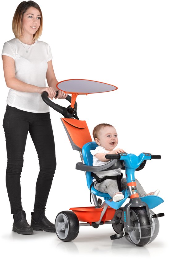 Vrouw duwt een blauwe driewieler met een baby erop. De driewieler heeft een oranje mandje en zonnekap.