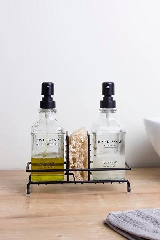 Glazen fles met 'HAND SOAP' erop, naast een fles met 'DISH SOAP' en een spons. Alles op een zwarte houder.