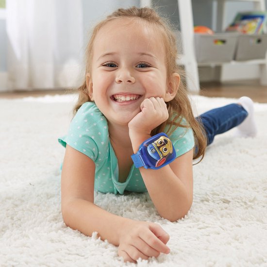 Een lachend meisje ligt op een kleed en draagt een blauw Paw Patrol horloge met Chase erop.