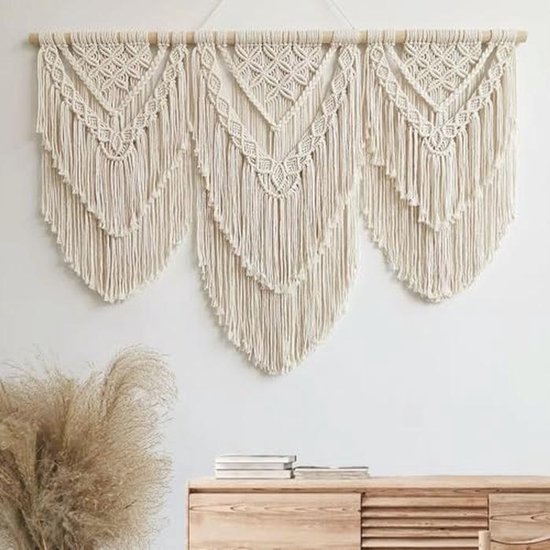 Crèmekleurig macramé wandkleed met drie V-vormige patronen, boven een houten kastje en siergrassen.