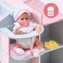 Pop met roze slab en muts, zittend in de kinderstoel van een grijs poppencommode met roze details. Gele schoentjes ernaast.
