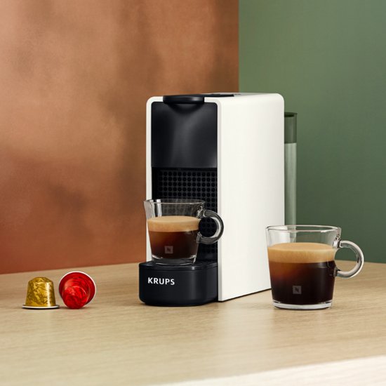 Machine à café Nespresso Essenza Mini blanche sur une table en bois, avec deux tasses d'expresso et des capsules.