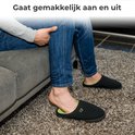 Man met spijkerbroek trekt een zwarte pantoffel met groene voering aan op een donkergrijs tapijt.