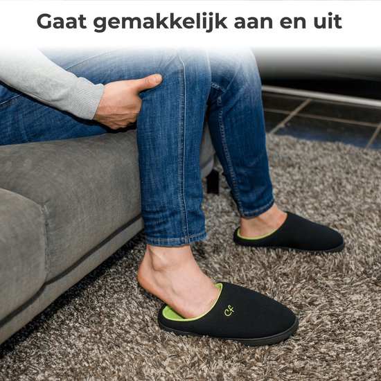 Man met spijkerbroek trekt een zwarte pantoffel met groene voering aan op een donkergrijs tapijt.
