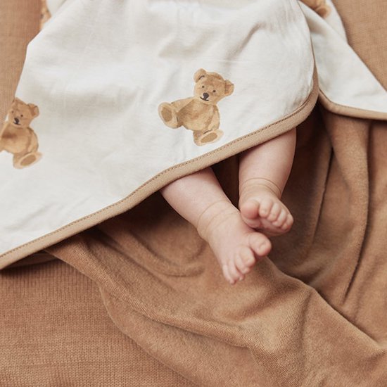 Un bébé aux pieds nus sous une cape de bain marron clair à imprimé oursons. La cape de bain a un bord beige.