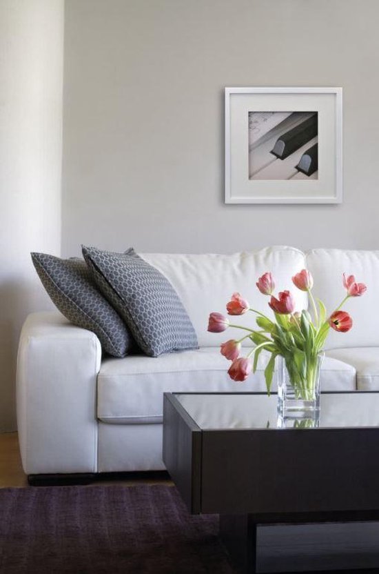 Een woonkamer met een witte bank, twee grijze kussens, een bijzettafel met een vaas bloemen en een witte fotolijst aan de muur.