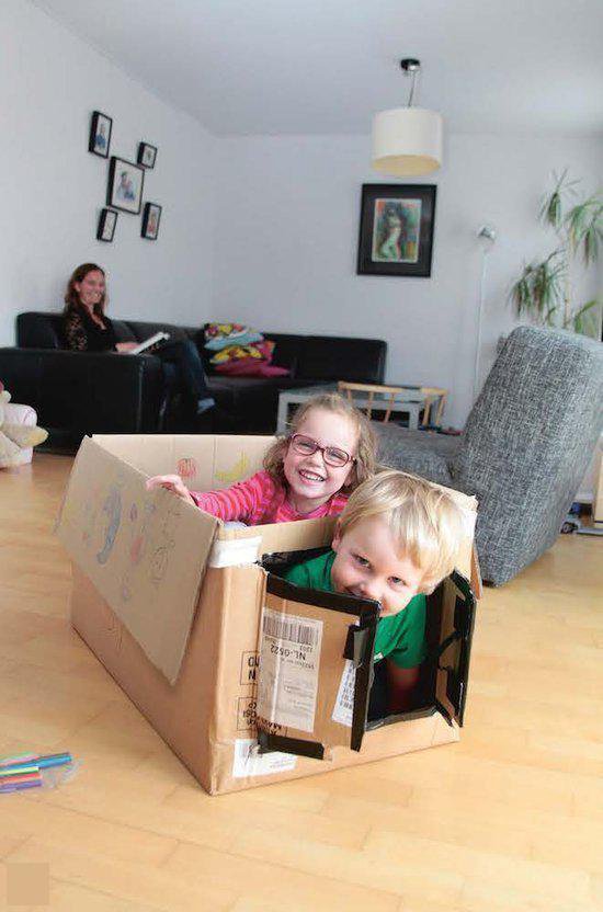 Twee lachende kinderen, een meisje met bril en een jongen, spelen in een kartonnen doos in een woonkamer.