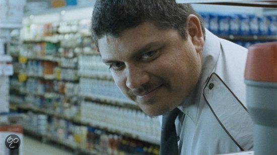 Close-up van een glimlachende man in een winkeluniform met achtergrond van schappen.
