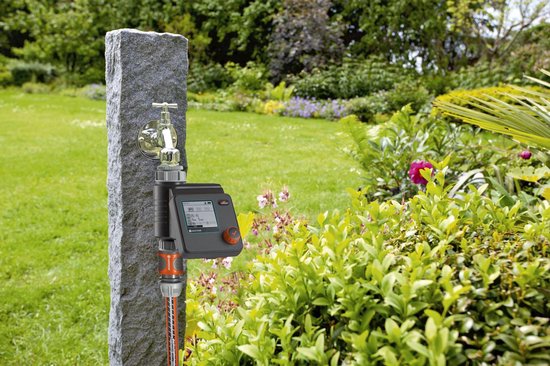 GARDENA besproeiingscomputer bij kraan op stenen pilaar. Groene tuin met bloemen.