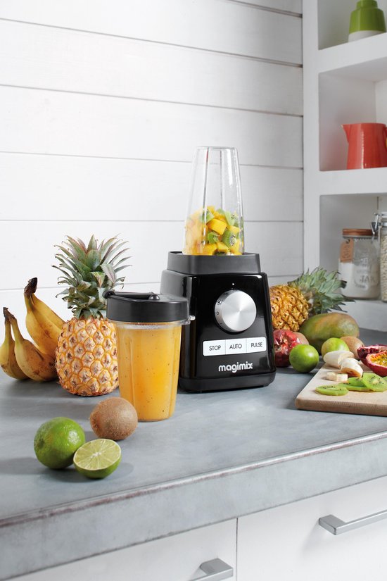 Zwarte Magimix blender met een beker vol stukjes fruit, omringd door vers fruit zoals ananas, bananen en kiwi.