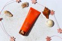 Een oranje Lancaster tube "Golden Tan Maximizer" ligt op wit zand, omringd door schelpen en roze bloemen.