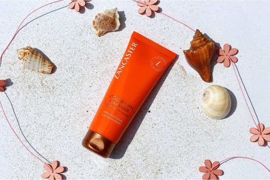 Een oranje Lancaster tube "Golden Tan Maximizer" ligt op wit zand, omringd door schelpen en roze bloemen.