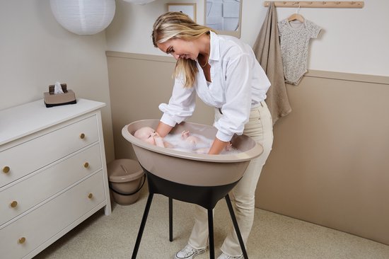 Een vrouw wast een baby in een beige Luma badje met zwart onderstel. Een commode en luiertafel staan ernaast.