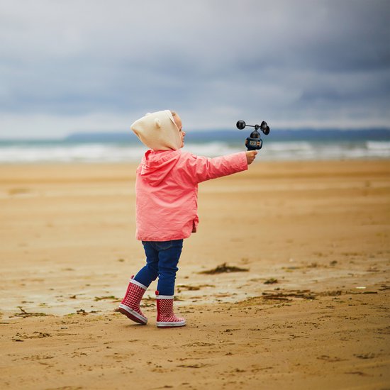 Kind met roze jas en laarsjes op strand houdt weerstation vast, met zee op achtergrond.