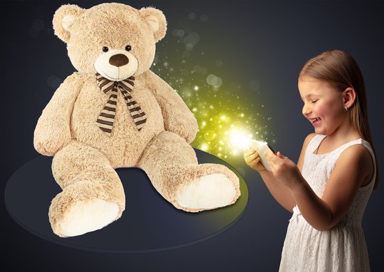 Een grote beige teddybeer kijkt naar een lachend meisje in een witte jurk dat een mobiele telefoon vasthoudt.