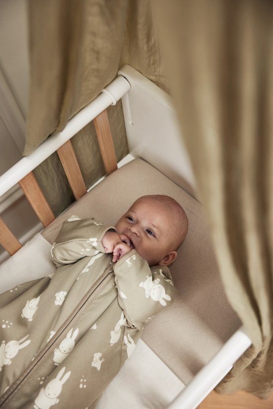 Baby in een olijfgroene slaapzak met Miffy print, liggend in een ledikant.
